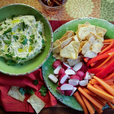 a bowl camellia bran beans hummas next to a plate of pita bread, carrots, and orange bell peppers