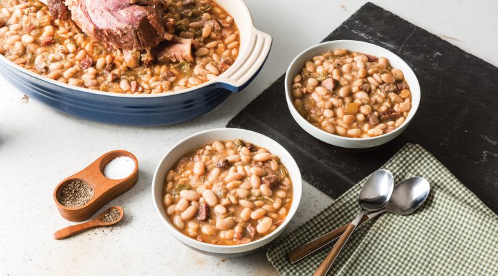 a close up of a roaster pot with white beans and a hamhock and 2 bowls full of the beans