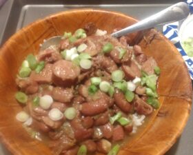 A wooden bowl filled with red beans, sliced sausage, and chopped green onions served over white rice, with a spoon in the bowl. In the background are tomatoes, an apple, and other food items.