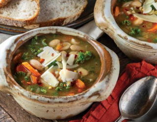 Two rustic bowls of hearty bean and vegetable soup garnished with shaved cheese and red pepper flakes. A spoon rests nearby on a red cloth, and crusty bread slices can be seen in the background.