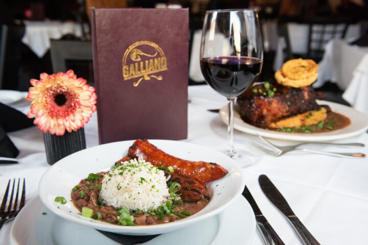 Table, with white tablecloth, at Galliano's Restaurant, with plated Pork Chop LaPlace entree, glass of red wine, to right, and their menu standing up behind plate