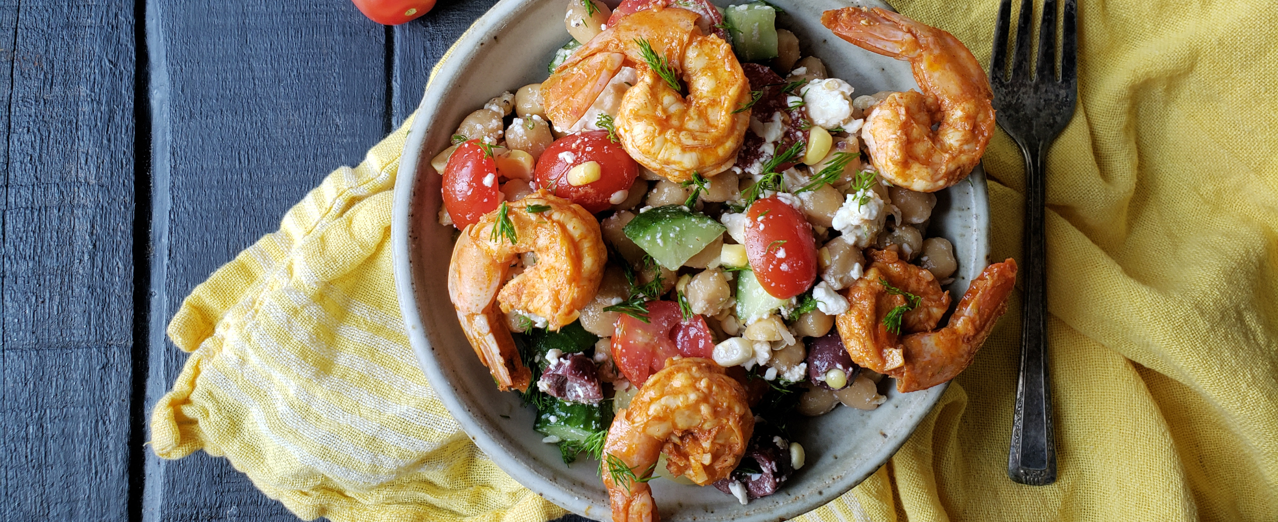 A bowl of shrimp salad with cherry tomatoes, cucumbers, olives, and feta on a yellow cloth. The setting is rustic with a dark wood table.
