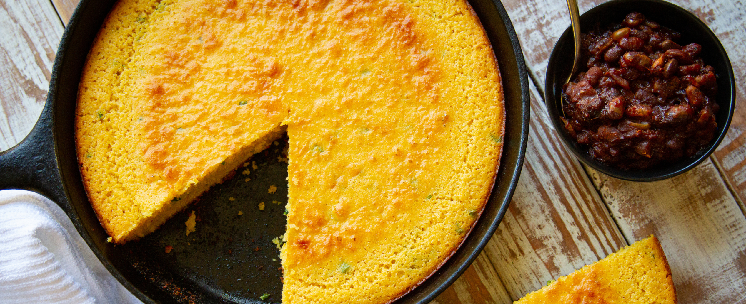 A golden cornbread in a cast iron skillet has a wedge cut out. A knife rests on the edge. A bowl of baked beans is beside the skillet on a rustic table.