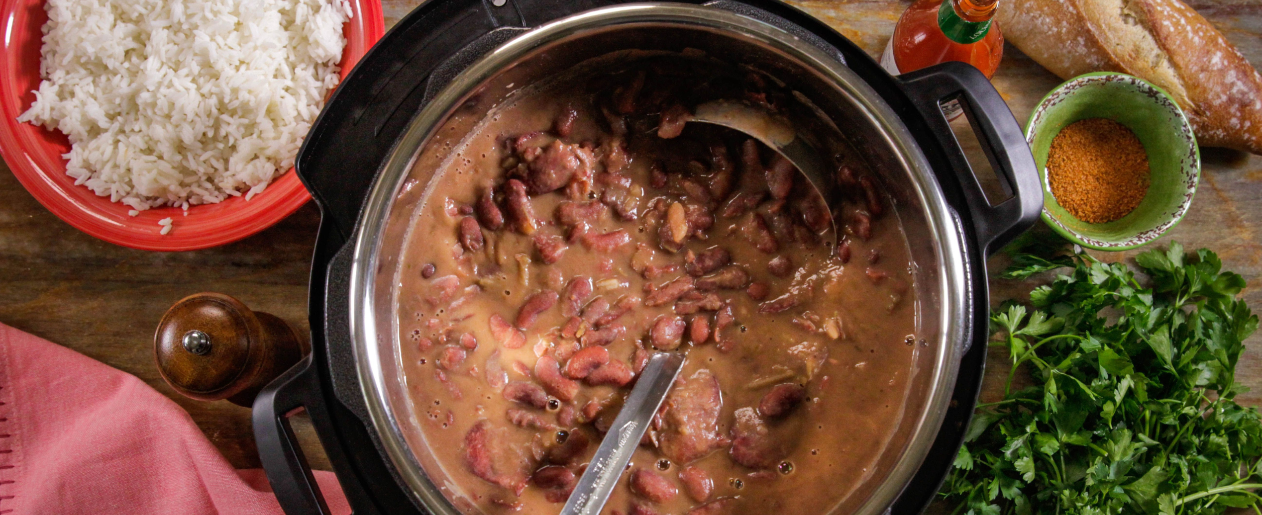 A pot of red beans with a ladle sits on a wooden table, surrounded by cooked rice, a baguette, spice bowl, fresh parsley, hot sauce, and a pepper mill.