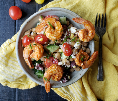 A bowl of shrimp salad with cherry tomatoes, cucumbers, olives, and feta on a yellow cloth. The setting is rustic with a dark wood table.