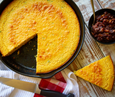 A golden cornbread in a cast iron skillet has a wedge cut out. A knife rests on the edge. A bowl of baked beans is beside the skillet on a rustic table.