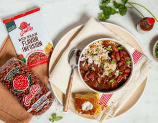 Bowl of red beans and rice with green onions on a napkin-lined plate, next to cornbread with butter, Camellia beans and flavor mix, hot sauce, and herbs.