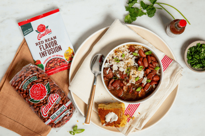 Bowl of red beans and rice with green onions on a napkin-lined plate, next to cornbread with butter, Camellia beans and flavor mix, hot sauce, and herbs.