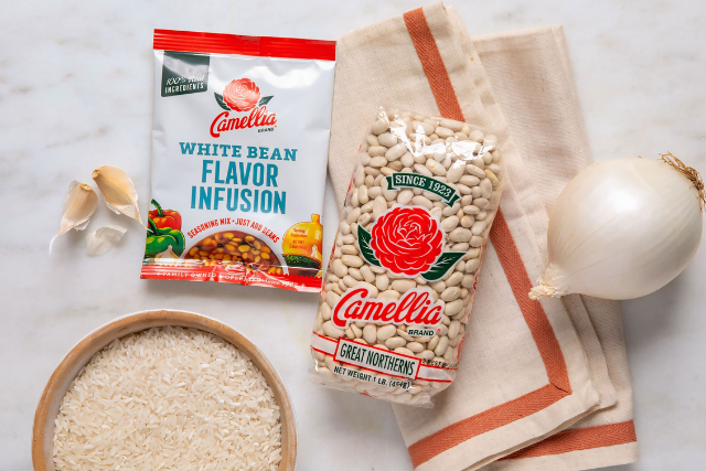 Package of Camellia white beans and seasoning mix on a marble surface, surrounded by garlic cloves, an onion, rice, and a beige kitchen towel.