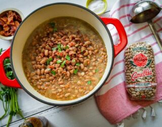 overhead image of large, red, pot of cooked pinto beans dish, with 1 pound bag of Camellia Pinto dry beans to its right, on table with a striped napkin and silver ladel