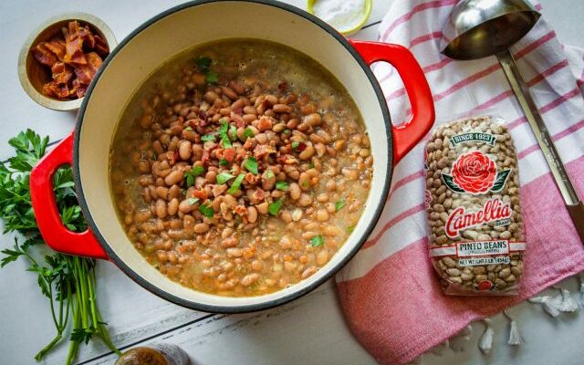 overhead image of large, red, pot of cooked pinto beans dish, with 1 pound bag of Camellia Pinto dry beans to its right, on table with a striped napkin and silver ladel