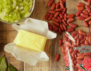 Stick of butter partially unwrapped on wood table, next to chopped green celery, scattered red beans, and a bag of packaged red beans. Rustic cooking theme.
