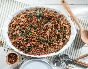 A white oval dish filled with cooked black-eyed peas and greens on a striped cloth. Nearby are a wooden spoon, utensils, and a bowl of red pepper flakes.