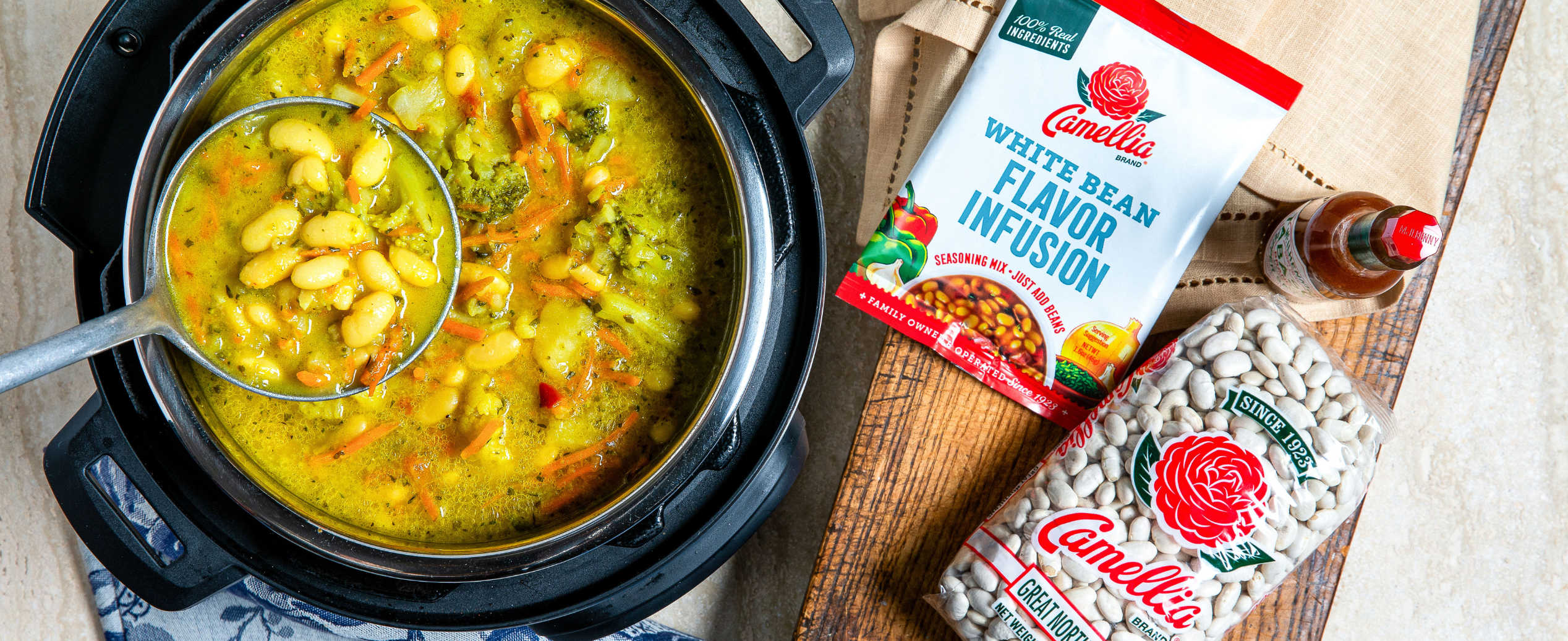 An inviting close-up of a pot of white bean stew with colorful vegetables and herbs. Nearby are Camellia bean packages and a bottle of hot sauce on a wooden board.