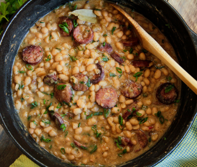 A hearty stew of white beans and sliced sausage simmers in a black pot, garnished with fresh herbs and a bay leaf. A wooden spoon rests on the side.