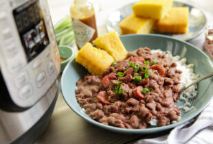 A bowl of red beans and rice topped with green onions and sausage slices, served with cornbread. Nearby are a pressure cooker and hot sauce bottle.