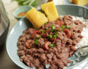 A bowl of red beans with sausage slices and green onions over white rice, accompanied by cornbread pieces. The setting is warm and homely.
