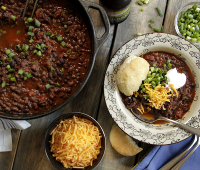 A rustic table setting features a pot of chili topped with green onions, a bowl of grated cheese, and a plate of chili with toppings, a roll, and a napkin.