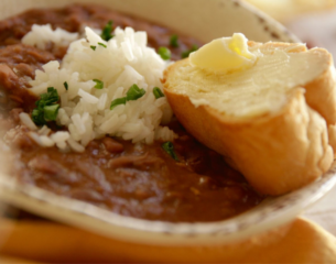 A bowl of savory red beans topped with white rice and chopped green herbs, served alongside a slice of bread spread with butter. Warm and comforting.