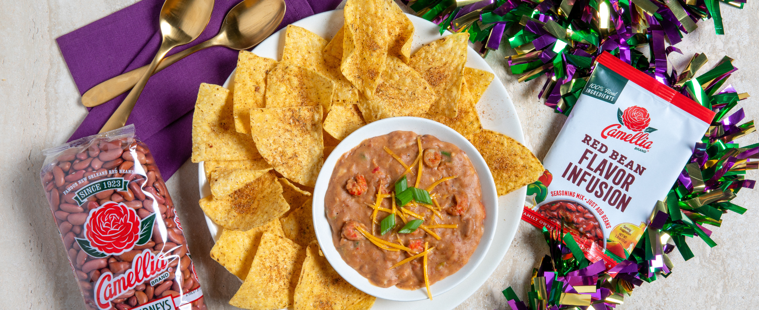 Plate of seasoned tortilla chips with a bowl of red bean dip garnished with cheese and herbs. Bags of beans and festive decorations are nearby. Colorful and festive.