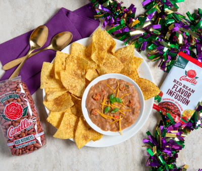 A bowl of bean dip garnished with green herbs sits on a plate surrounded by tortilla chips. A bag of red kidney beans, a flavor packet, two gold spoons, a purple napkin, and colorful beads are arranged nearby, creating a festive atmosphere.