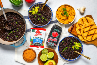 A colorful spread featuring black beans in bowls, a pot, and packets, alongside grilled tortillas, cilantro rice, lime slices, spices, and garlic.