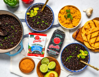 Overhead shot of a table with a pot and 2 bowls of cooked, stovetop chipotle black beans, along with 1 package of chipotle black beans flavor infusion, one, 1-pound bag of black beans, a small bowl of limes and an avocado, some spices, a garlic clove, two bell peppers, a bowl of cooked rice, and some grilled tortillas that are on a small wooden cutting board.