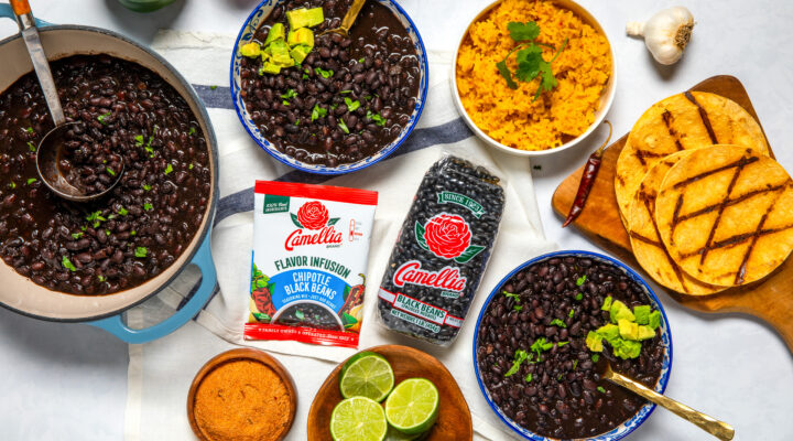 Overhead shot of a table with a pot and 2 bowls of cooked, stovetop chipotle black beans, along with 1 package of chipotle black beans flavor infusion, one, 1-pound bag of black beans, a small bowl of limes and an avocado, some spices, a garlic clove, two bell peppers, a bowl of cooked rice, and some grilled tortillas that are on a small wooden cutting board.