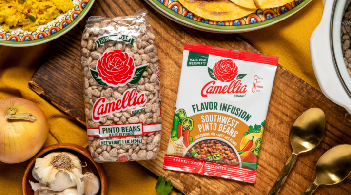 Camellia Pinto Beans and Southwest Pinto Beans Flavor Infusion on a wooden board with garlic, onion, and herbs, alongside cooked rice, tortillas, and a pot of beans.