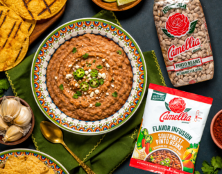 Camellia Pinto Beans and Southwest Pinto Beans Flavor Infusion on a wooden board with garlic, onion, and herbs, alongside refried pinto beans and tortillas.