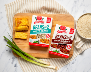 Boxes of Camellia white and red beans sit on a wooden board with cornbread squares and green onions. A bowl of rice is nearby on a striped cloth.