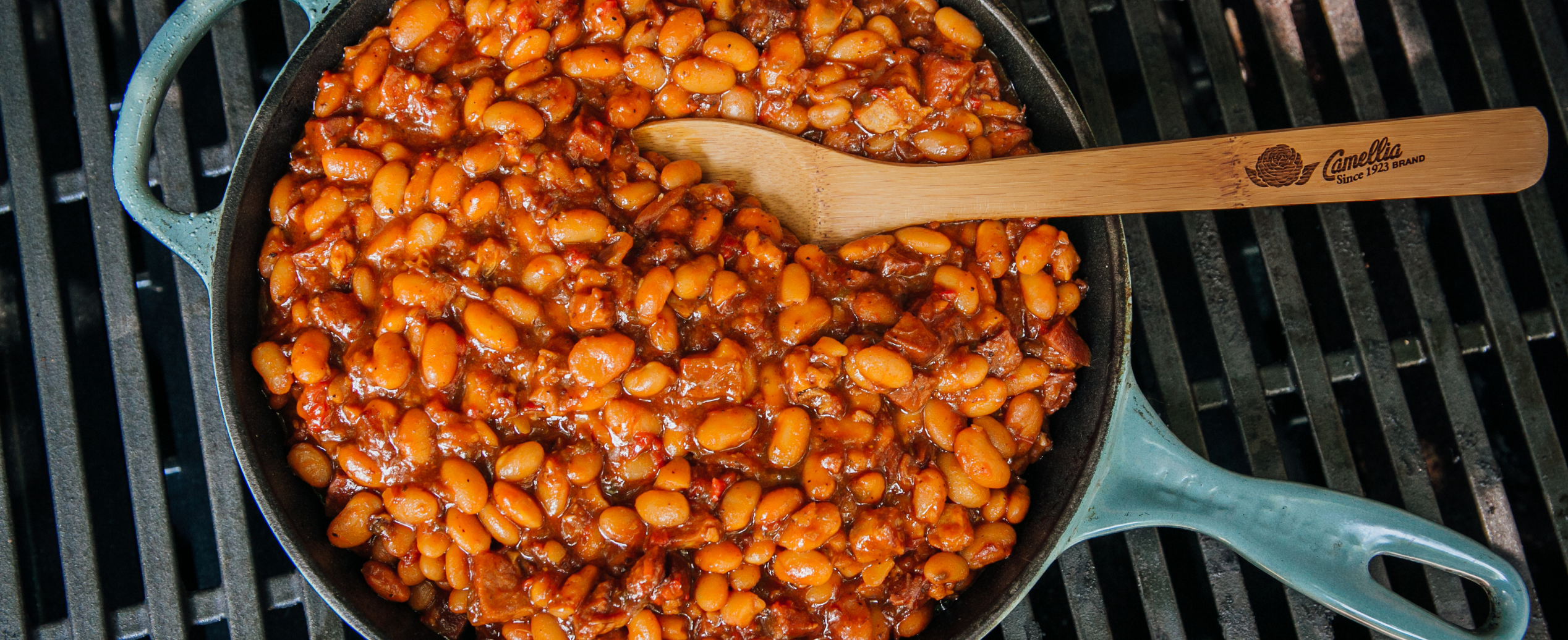 Cast-iron skillet with baked beans, featuring a rich sauce and bacon pieces, sits on a barbecue grill. A wooden spoon rests inside, evoking a hearty, home-cooked feel.