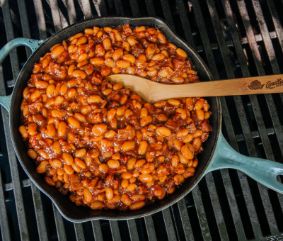 Cast-iron skillet with baked beans, featuring a rich sauce and bacon pieces, sits on a barbecue grill. A wooden spoon rests inside, evoking a home-cooked feel.
